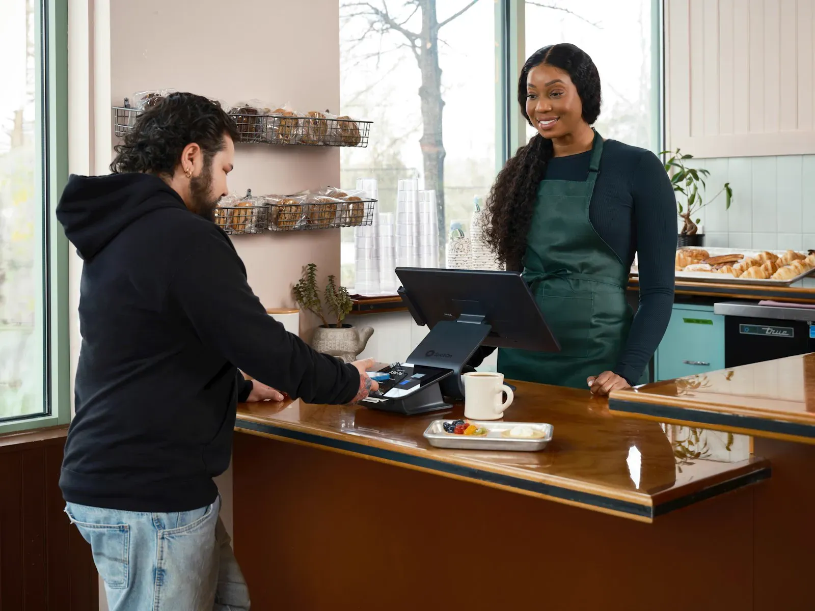 Customer paying with a smartphone at a cafe counter while a barista in a green apron smiles, a typical post-service moment when a review request would land best.