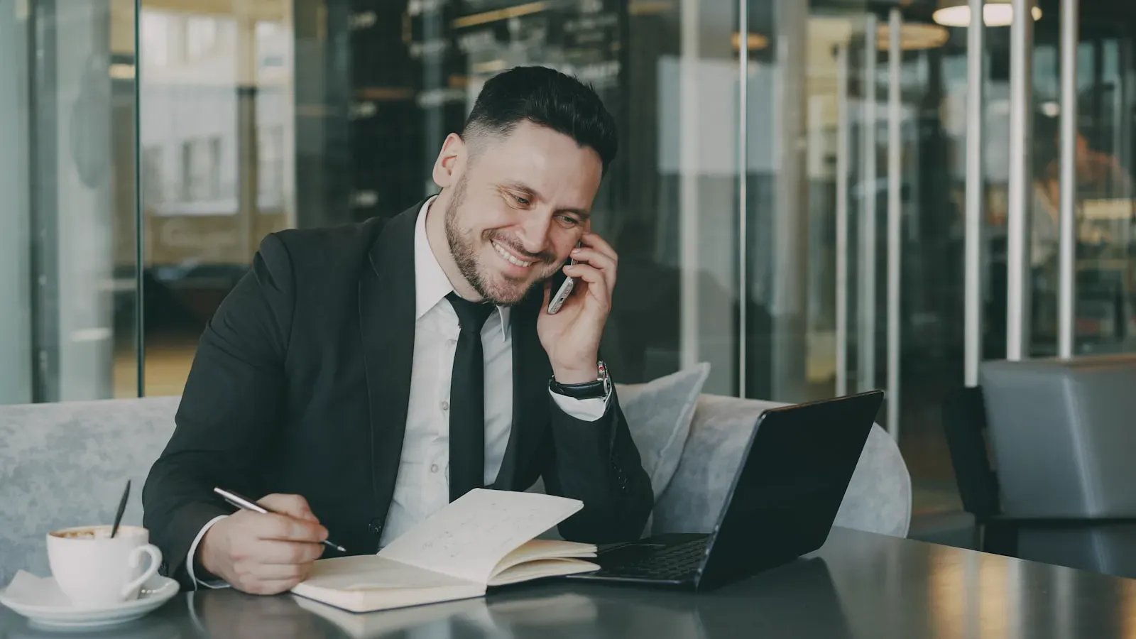 Businessman in professional attire taking notes while on a phone call, capturing the moment of a reference check or vendor interview.