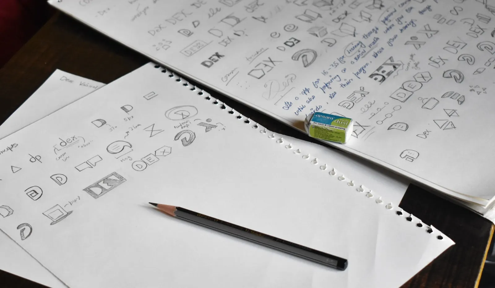 A pencil resting on top of a pile of paper showing logo design sketches, representing the early-stage exploration that precedes a finished brand mark.