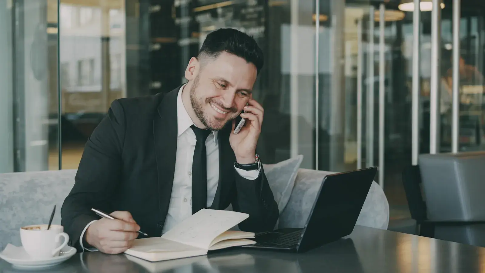 Businessman in professional attire taking notes while on a phone call, capturing the moment of a reference check or vendor interview.